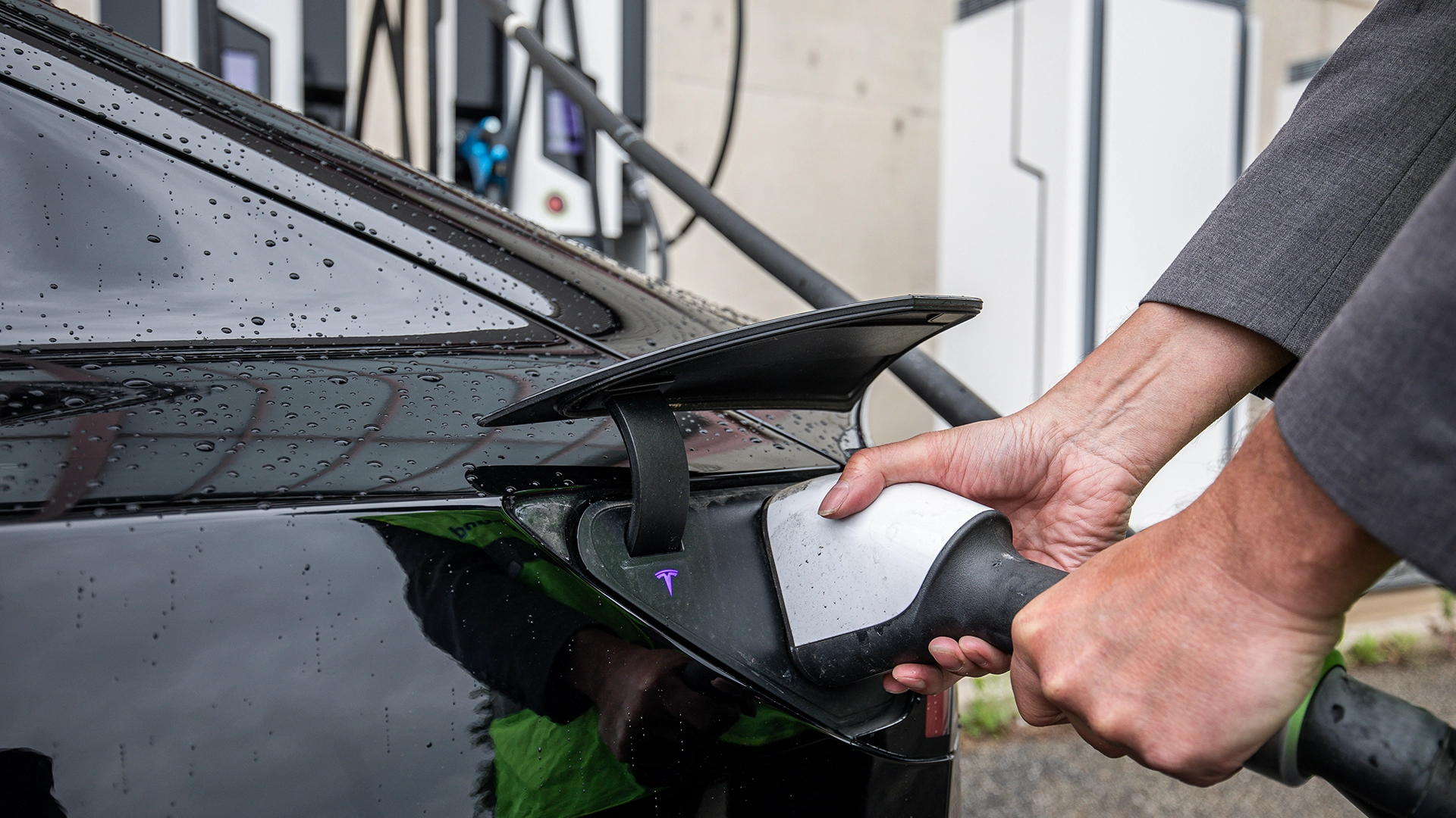 The image shows the charging process of an electric vehicle as part of the E-VIA FLEX-E project. A person is connecting a high-power charging cable to the charging port of a black electric vehicle. High-power charging stations are visible in the background.